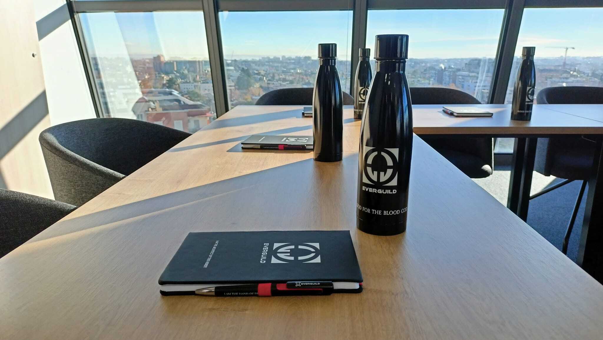 Everguild business merchandising There are a few bottles and a notebook in colour black with the Everguild logo printed on them, white over black. These things are laying on a table in a bright office room.